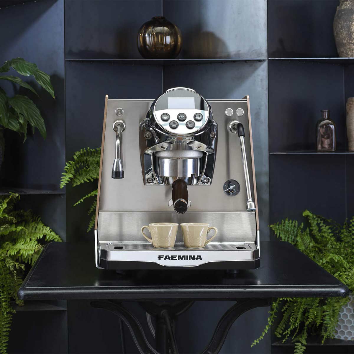 A silver Faema Faemina Dual Boiler Espresso Machine with AUTOSTEAM sits on a black table, brewing two espresso shots into cups amid green plants and decorative items—a beautiful addition to any space. © 2025 1st-line.com