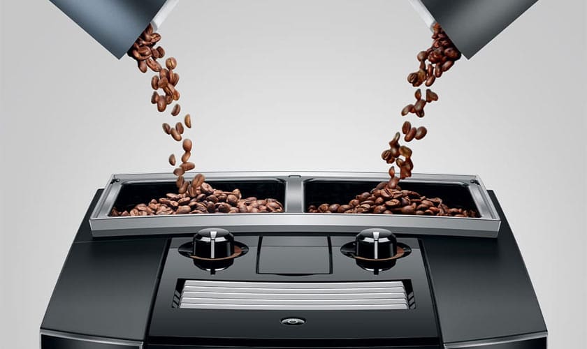 Close-up of two compartments on the Jura J8 Twin Super-Automatic Espresso Machine, each being filled with coffee beans pouring down. The modern design showcases sleek black and silver surfaces. © 2025 1st-line.com