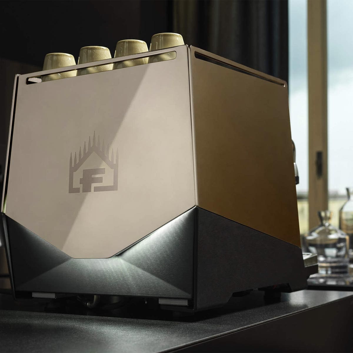 The Faema Faemina Dual Boiler Espresso Machine with AUTOSTEAM, featuring a modern metallic geometric design and an engraved logo, sits on a dark countertop near a window, with six cups on top and glassware in the background. © 2025 1st-line.com