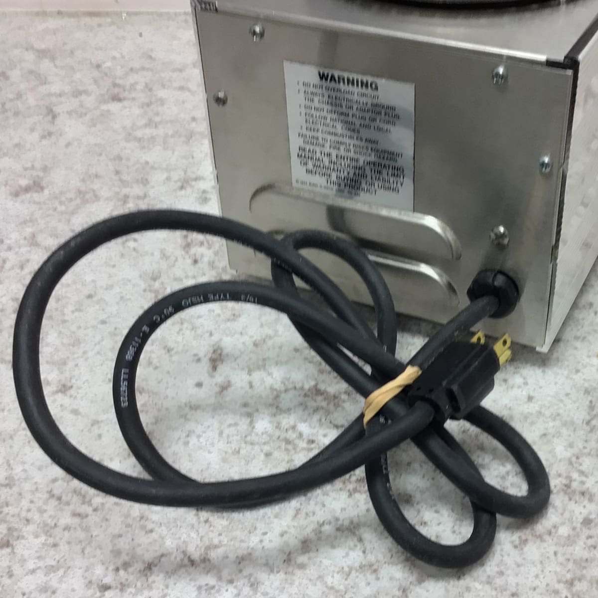 A black power cord is plugged into a SIDEWALK SALE - Bunn Dual Warmer (E109) on a light countertop. A warning label is visible on the back, and the cord is loosely coiled and secured with a rubber band. © 2025 1st-line.com