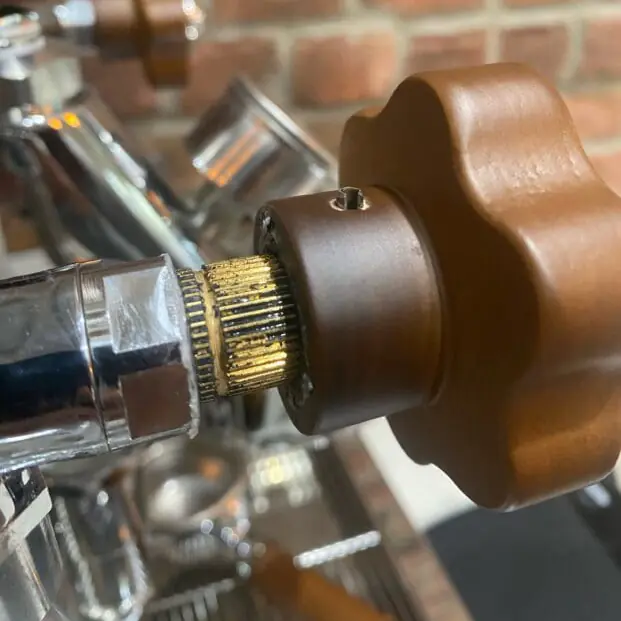 A close-up of a Lelit Bianca coffee machine&rsquo;s steam valve, featuring a brown, ridged knob and metallic components, with a background of blurred brick wall.