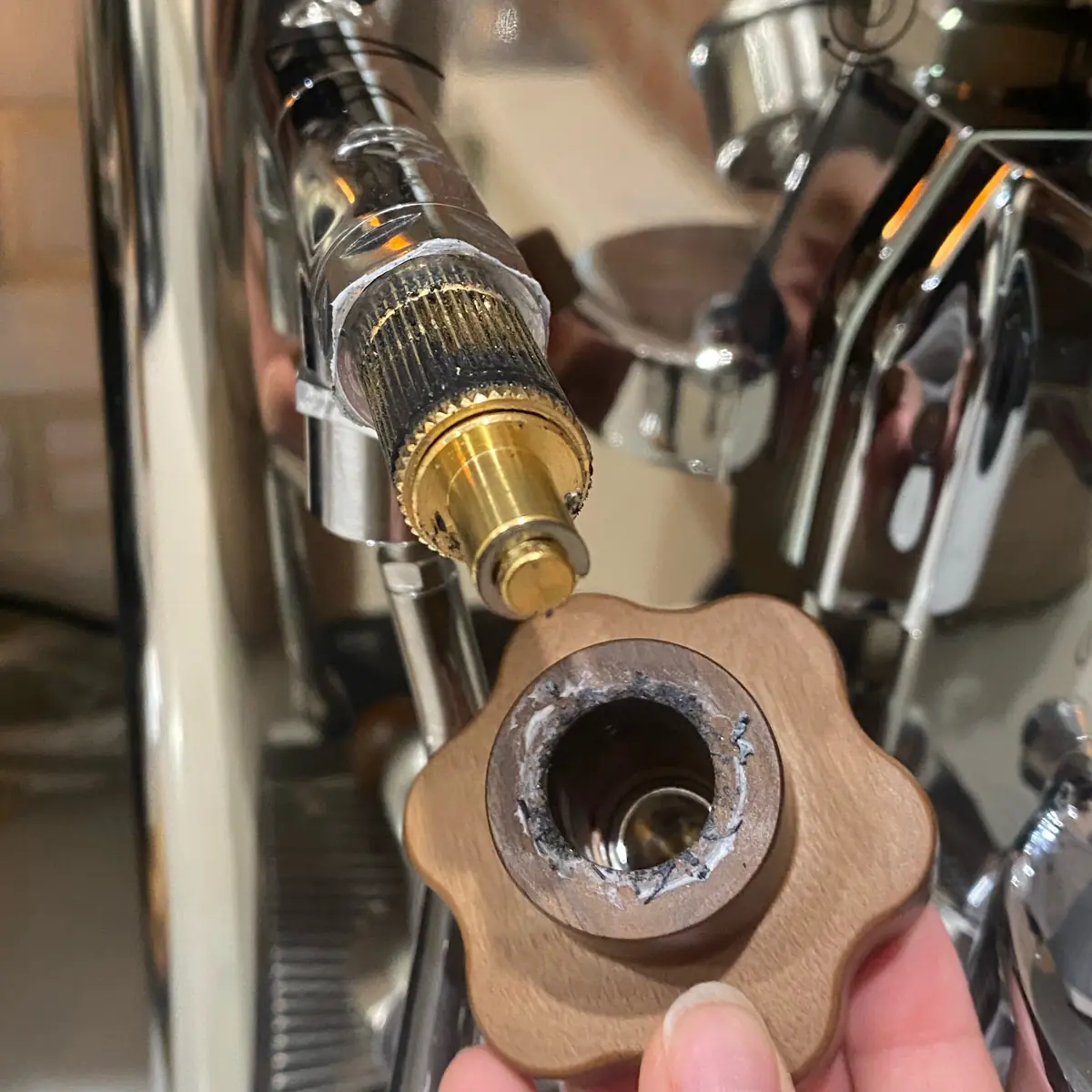 A hand holds a broken brown metal knob in front of a shiny Lelit Bianca espresso machine, showing the exposed gears where the steam valve knob has detached from the machine.