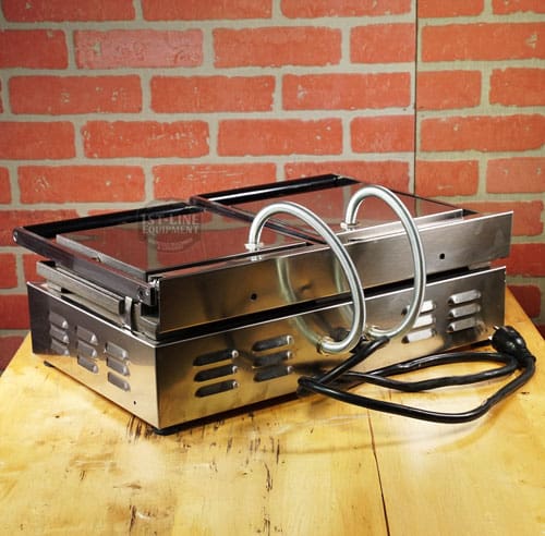 A SIDEWALK SALE - Sirman Panini Double Grill (E112) with glass lid, heating coils, and power cord sits on a wooden table before a red brick wall—ideal for your next sidewalk sale. © 2025 1st-line.com