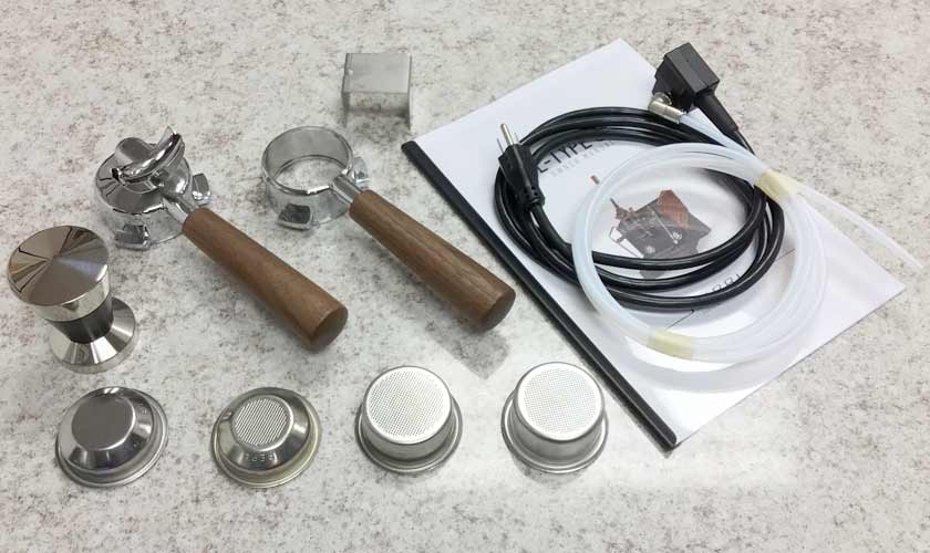 Accessories for the Nurri L-Type Lever Dual Boiler Espresso Machine on a countertop, including a portafilter, tamper, baskets, power cord, tubing, metal dosing tool, and user manual. © 2025 1st-line.com