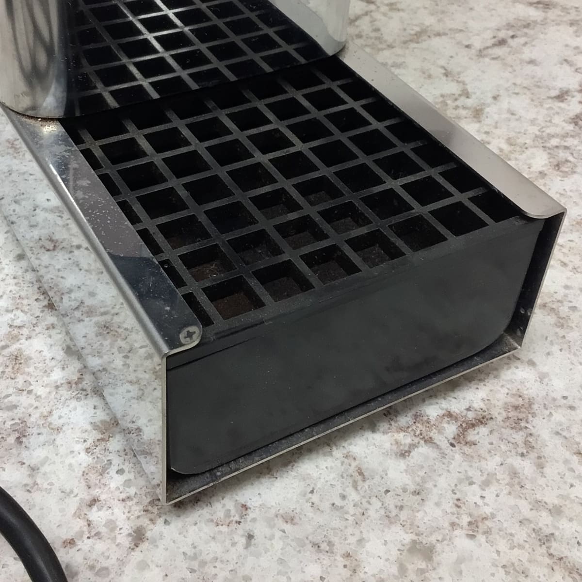 Close-up of a metal espresso machine drip tray with black grid on a speckled countertop. Nearby is the SIDEWALK SALE - Test Conical Burr Espresso Grinder (E109); a power cord is partially visible in the corner. © 2025 1st-line.com