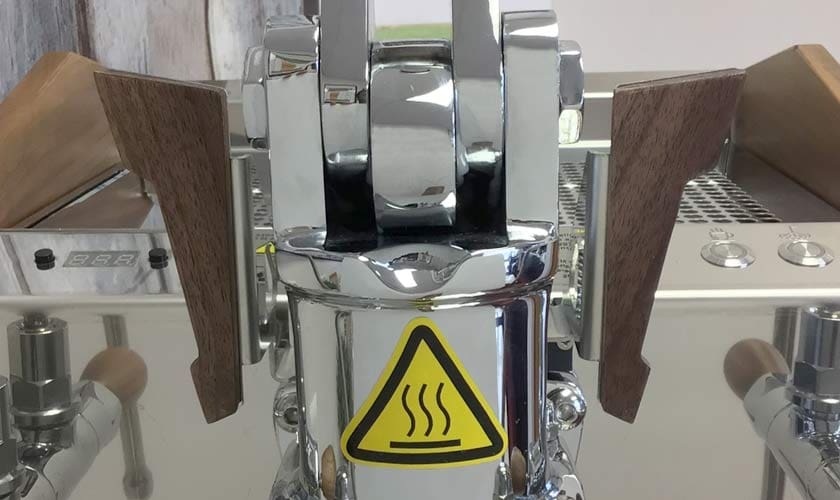 Close-up of the Nurri L-Type Lever Dual Boiler Espresso Machine, featuring shiny metal construction, wooden handles, and a yellow caution sign with three wavy heat lines indicating a hot surface. © 2025 1st-line.com