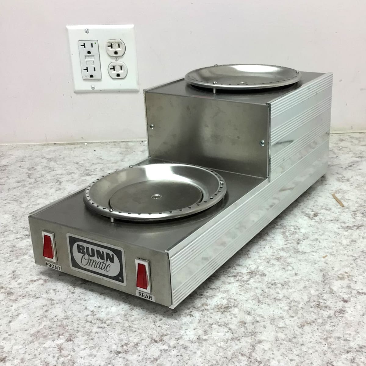 The SIDEWALK SALE - Bunn Dual Warmer (E109) with front and rear warming plates sits on a speckled countertop near a wall outlet—ideal for keeping coffee hot at your next sidewalk sale. © 2025 1st-line.com