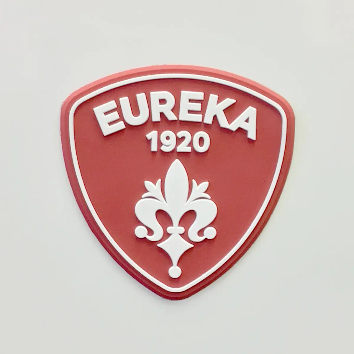 A red shield-shaped emblem with a white border, EUREKA at the top, 1920 below, and a stylized white fleur-de-lis in the center—mirroring the design featured on the Eureka Atom W 65 Weight Based Espresso Coffee Grinder. © 2025 1st-line.com