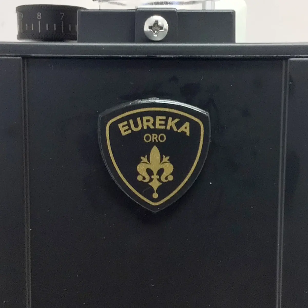 Close-up of a black Eureka Mignon Single Dose Espresso Coffee Grinder, showcasing a gold and black shield-shaped badge reading EUREKA ORO with a fleur-de-lis, and a numbered dial at the top left for advanced grind settings.