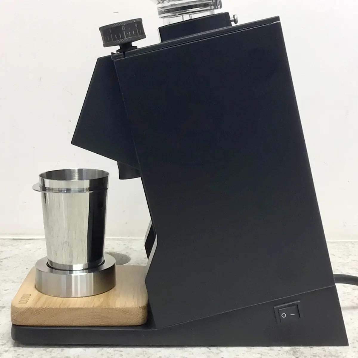 A Eureka Mignon Single Dose Espresso Coffee Grinder in sleek black sits on a counter with a metal cup under the dispenser, placed on a wooden platform. The on/off switch is visible at the bottom right of the machine.