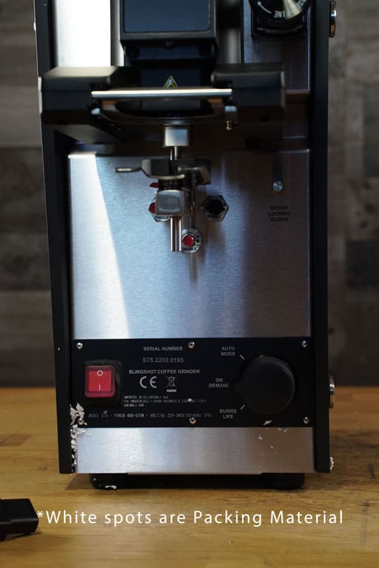 A close-up of a Slingshot Dosis S75 Espresso Coffee Grinder Black 220V shows white packing material attached to the bottom. Text below reads: *White spots are Packing Material. © 2025 1st-line.com