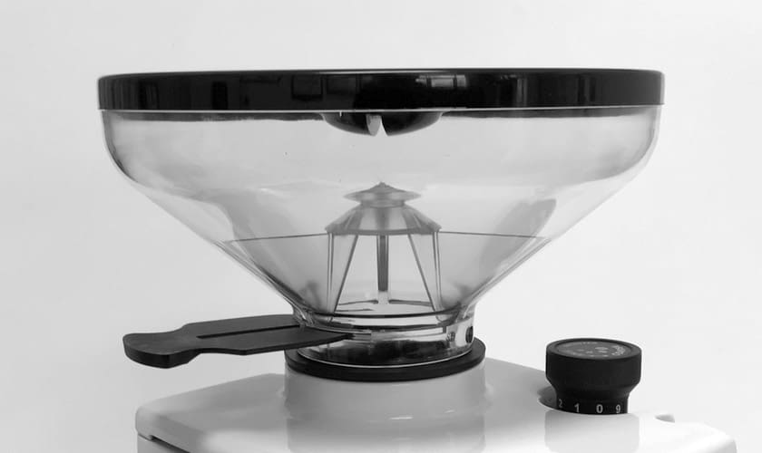 A close-up of the Eureka Atom W 65 Weight Based Espresso Coffee Grinder shows its transparent hopper with black lid on a white base. A numbered dial is visible on the right side of the grinder. © 2025 1st-line.com