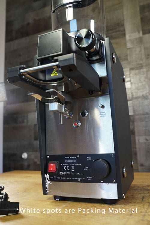 A close-up of the Slingshot Dosis S75 Espresso Coffee Grinder Black 220V on a wooden surface displays its buttons, dials, and portafilter holder. White spots near the base are identified as packing material. © 2025 1st-line.com
