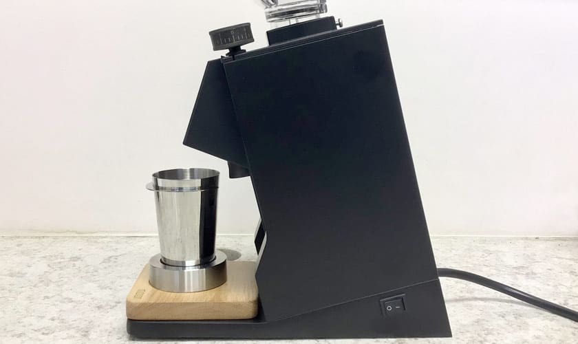 A Eureka Mignon Single Dose Espresso Coffee Grinder in black with a metal container and wooden base is placed on a light countertop, featuring a side power switch and top dial, set against a plain white wall. © 2025 1st-line.com
