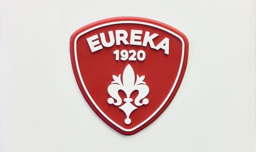 A red shield-shaped emblem with EUREKA in white above 1920 and a fleur-de-lis below, outlined in white, represents the quality of the Eureka Atom W 65 Weight Based Espresso Coffee Grinder. © 2025 1st-line.com