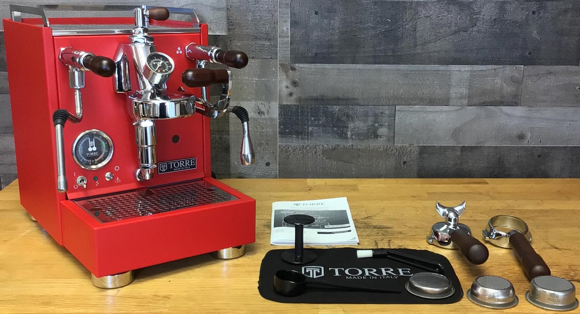 A red Torre Luigino TSP HX Espresso Machine w/ Flow Control sits on a wooden counter with portafilters, tamper, black mat, and manual—ideal for practicing Flow Control techniques against a rustic wood-paneled backdrop. © 2025 1st-line.com