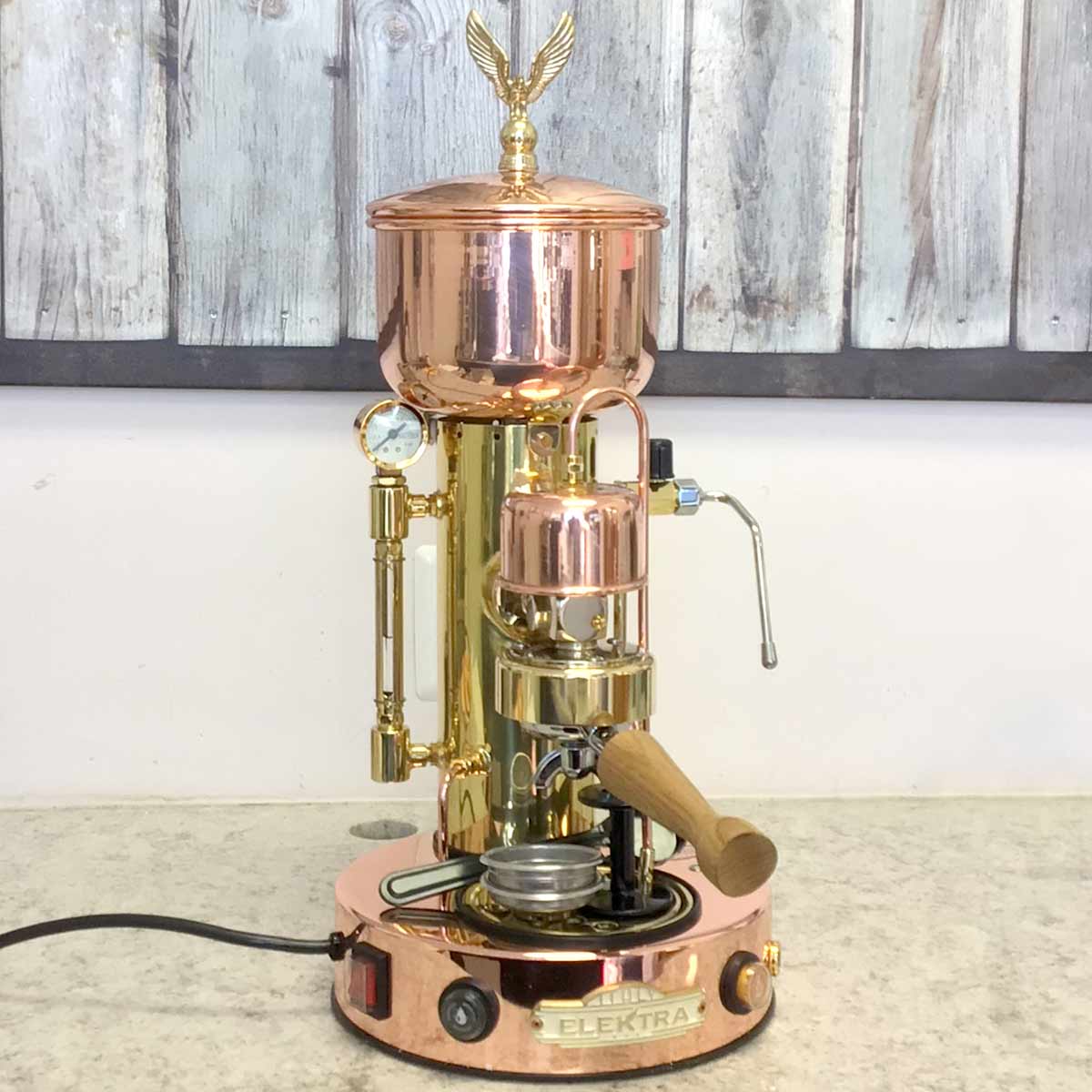 A returned, refurbished Elektra SX Microcasa Semiautomatica Espresso Machine (E210) from the SIDEWALK SALE sits on a countertop, showcasing a vintage style with polished copper and brass accents plus a wooden-handled portafilter. © 2025 1st-line.com