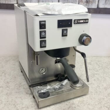 A SIDEWALK SALE - Rancilio Silvia Pro X White Dual Boiler Espresso Machine (CONVENTION DEMO) (E113) sits on a countertop with visible buttons, steam wand, portafilter attached, and a wrapped accessory on the drip tray. © 2025 1st-line.com