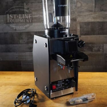 A Slingshot Dosis S75 Espresso Coffee Grinder Black 220V rests on a wooden surface beside its power cord and packaging, featuring 1ST-LINE EQUIPMENT branding. Note: White spots in the image are visible packing material. © 2025 1st-line.com