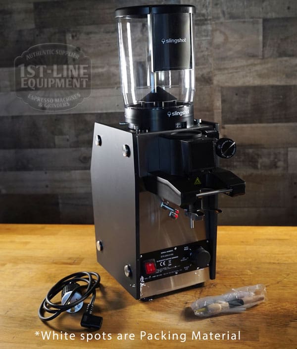 A Slingshot Dosis S75 Espresso Coffee Grinder Black 220V rests on a wooden surface beside its power cord and packaging, featuring 1ST-LINE EQUIPMENT branding. Note: White spots in the image are visible packing material. © 2025 1st-line.com