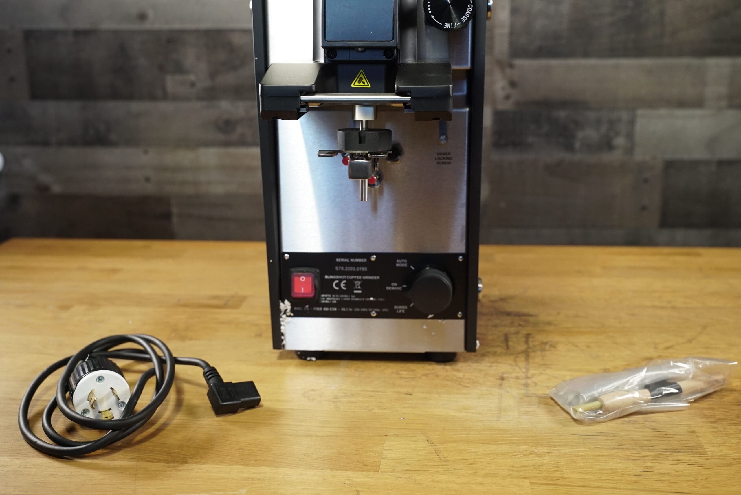 A Slingshot Dosis S75 Espresso Coffee Grinder Black 220V rests on a wooden surface with its power cable and parts bag in front, set against a classic wood-paneled wall backdrop. © 2025 1st-line.com