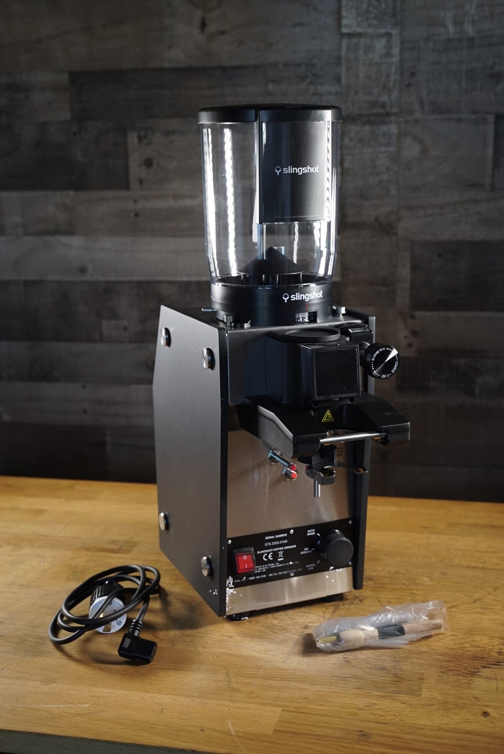 A Slingshot Dosis S75 Espresso Coffee Grinder Black 220V rests on a wooden surface with its power cord and a small brush in plastic packaging nearby. The scene is set against a dark, textured wooden wall. © 2025 1st-line.com