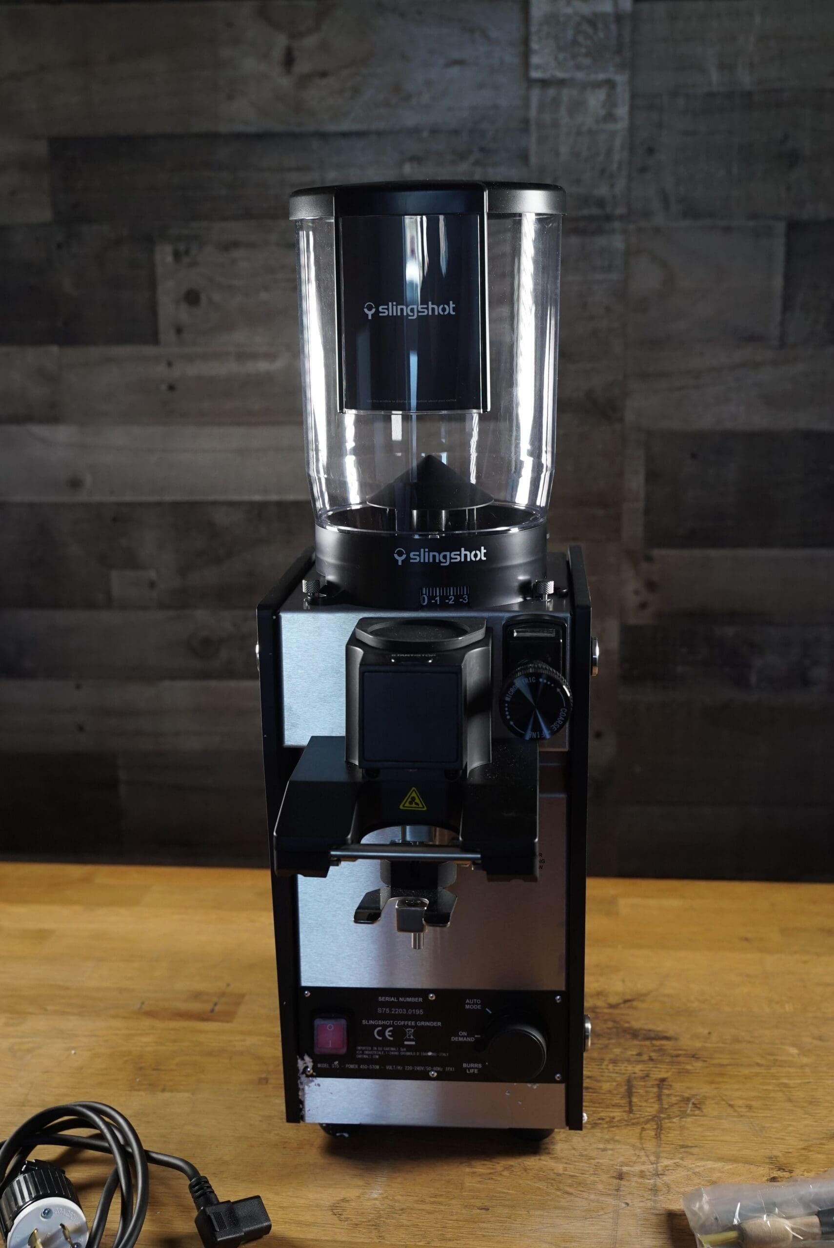A Slingshot Dosis S75 Espresso Coffee Grinder Black 220V sits on a wooden surface against a dark, textured wall, with its electric power cord placed nearby on the table. © 2025 1st-line.com