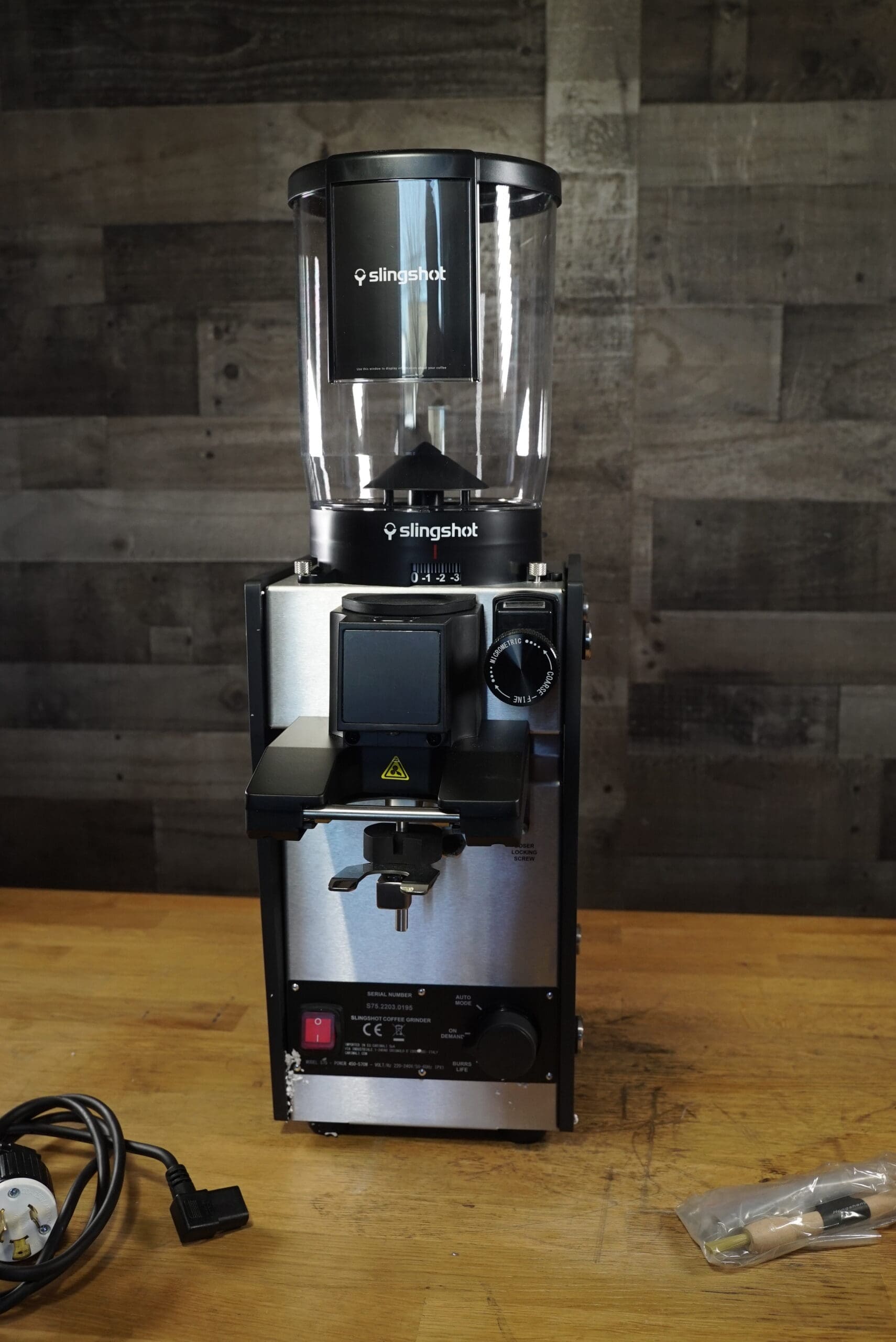 A Slingshot Dosis S75 Espresso Coffee Grinder Black 220V sits on a wooden surface near an unplugged power cord and a small bag with a metal tool, with a gray wooden wall in the background. © 2025 1st-line.com
