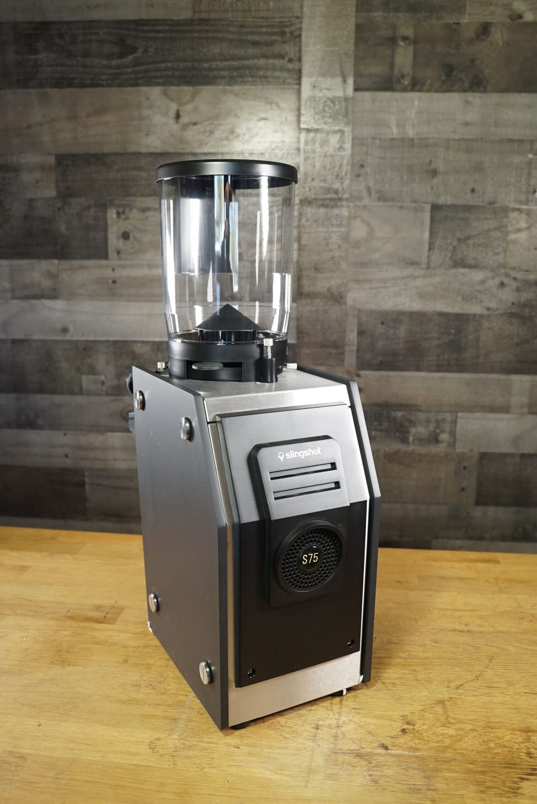 A Slingshot Dosis S75 Espresso Coffee Grinder Black 220V with a clear bean hopper sits on a wooden countertop, set against gray wooden paneling. © 2025 1st-line.com