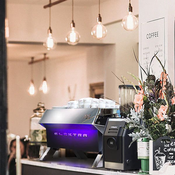 A stylish coffee shop counter features a CONVENTION DEMO USED 220 VOLTS Elektra Indie Smart 2GR 230V White/Stainless espresso machine, cups on top, a grinder, a vase of flowers, hanging bulbs, and a coffee menu on the wall. © 2025 1st-line.com