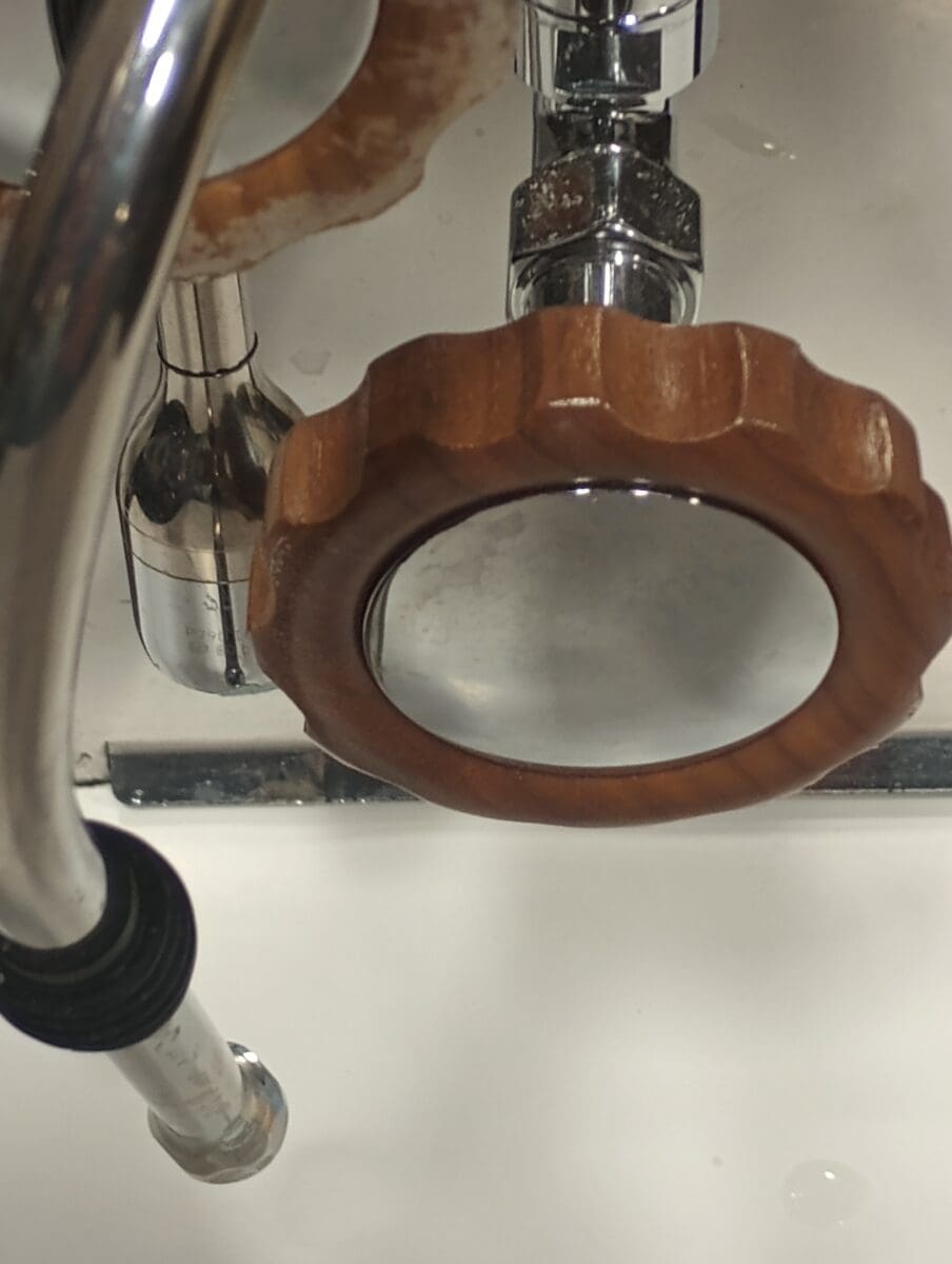 Close-up of an ACS Minima metal plumbing fixture with a large, round, grooved wooden valve handle above a white sink. Reflections and water spots are visible on the no steam, no hot water metal surfaces. © 2025 1st-line.com