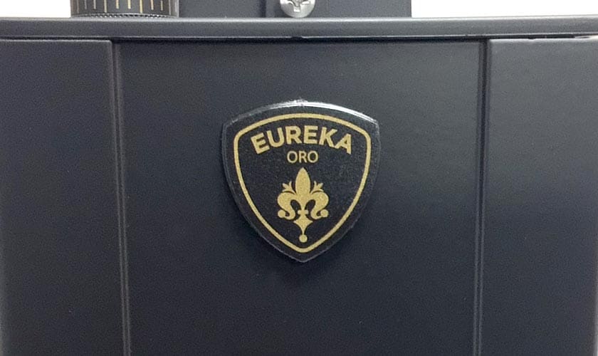 Close-up of the black Eureka Mignon Single Dose Espresso Coffee Grinder with a gold and black shield-shaped badge displaying EUREKA ORO and a fleur-de-lis—ideal for espresso enthusiasts. © 2025 1st-line.com