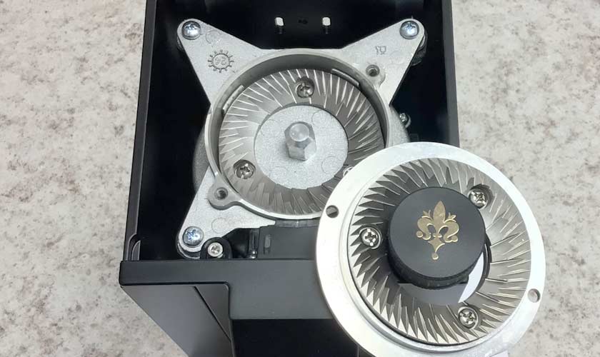 Close-up of the open Eureka Mignon Single Dose Espresso Coffee Grinder, revealing sharp metal burrs and intricate gear-like parts, with one burr cover removed and placed beside the grinder on a speckled countertop. © 2025 1st-line.com