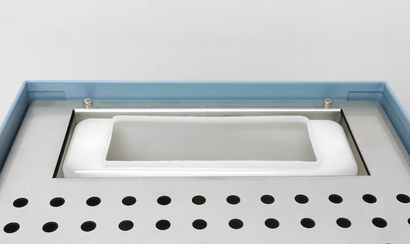 Close-up of the Torre Luigino TSP HX Espresso Machine with flow control, showing its rectangular white container set in a gray surface with round holes, under a transparent lid framed in blue. © 2025 1st-line.com