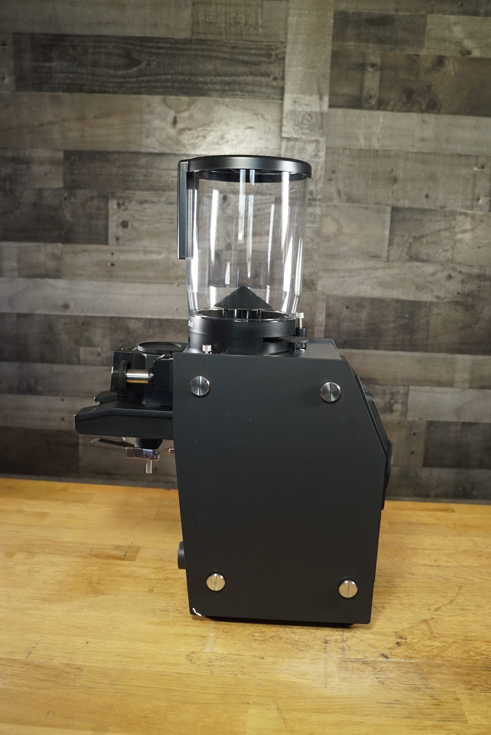 The Slingshot Dosis S75 Espresso Coffee Grinder Black 220V, featuring a transparent bean hopper, rests on a wooden countertop against a rustic wood panel wall—ideal for your 220V setup. © 2025 1st-line.com