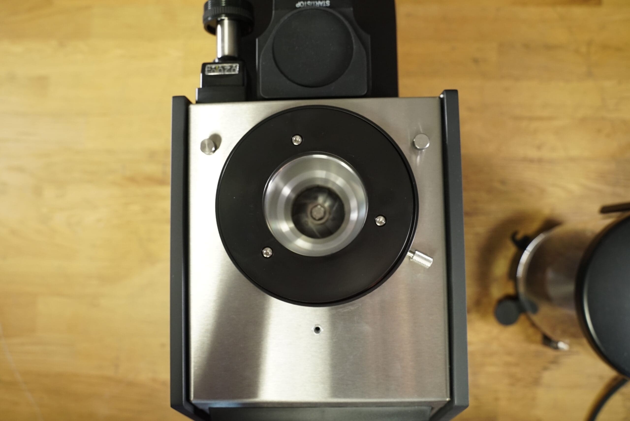 Top-down view of the Slingshot Dosis S75 Espresso Coffee Grinder Black 220V, with a metal and black finish and central grinding chamber on a wooden surface. Part of a kettle appears on the right. © 2025 1st-line.com