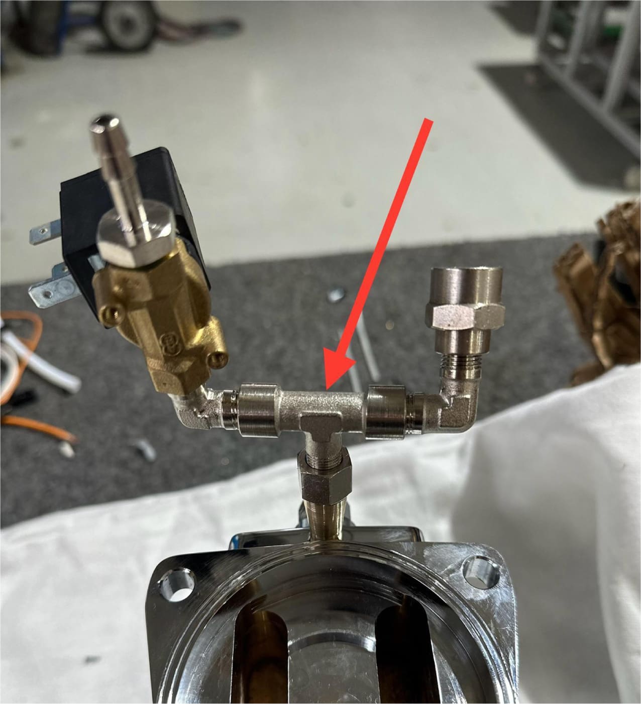 A close-up of the ACS Vostok's metal pipe assembly with various fittings and a solenoid valve, with a red arrow pointing to a T-shaped pipe joint at the center.