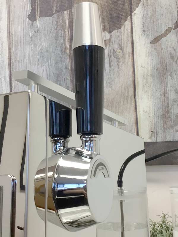 Close-up of a shiny Bellezza Bellona espresso machine portafilter with a black and silver handle, attached to a metallic coffee machine against a wooden background—the right fit for any coffee enthusiast. © 2025 1st-line.com