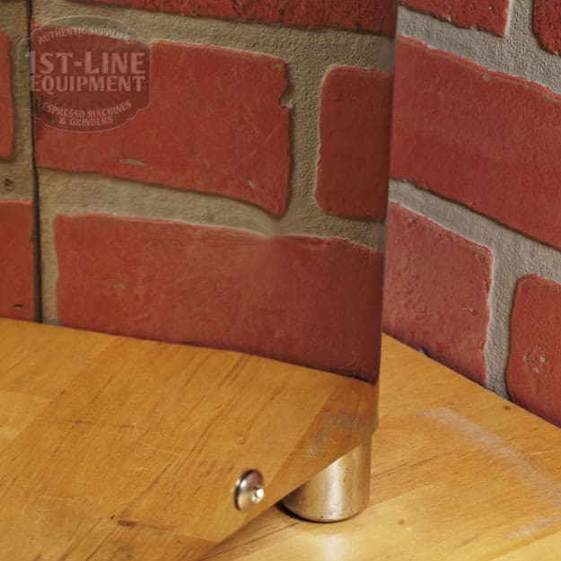 A chrome leg supports a wooden table in a room with red brick walls. Reflected in the leg are the bricks, tabletop, and a SIDEWALK SALE - VBM Domobar Super HX Tank Espresso Machine (USED) (E111) nearby. © 2025 1st-line.com