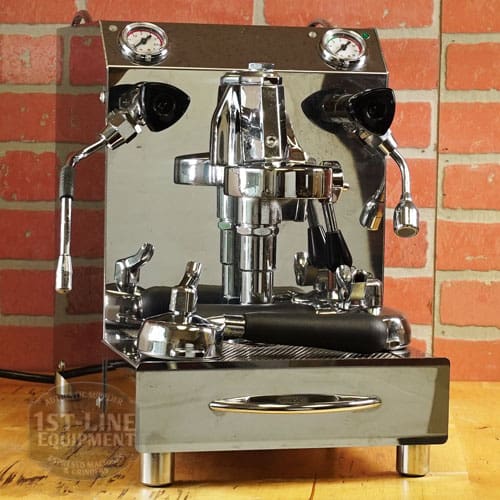 A shiny, used SIDEWALK SALE - VBM Domobar Super HX Tank Espresso Machine (E111) with dual gauges, steam wand, and portafilter rests on a wooden counter in front of a red brick wall. © 2025 1st-line.com