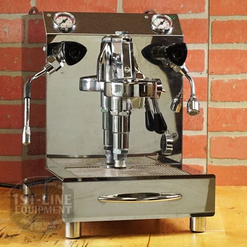 A shiny, used SIDEWALK SALE - VBM Domobar Super HX Tank Espresso Machine (E111) with two pressure gauges, steam wand, and hot water spout rests on a wooden surface in front of a red brick wall. © 2025 1st-line.com
