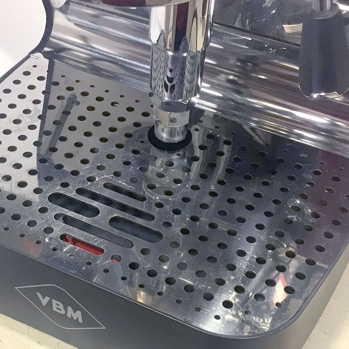 Close-up of the SIDEWALK SALE - VBM Super Digital DUAL PROFILER Dual Boiler Espresso Machine (used) (E111) drip tray with a metal grate reflecting machine parts above, and the portafilter head centered with water droplets on the tray. © 2025 1st-line.com