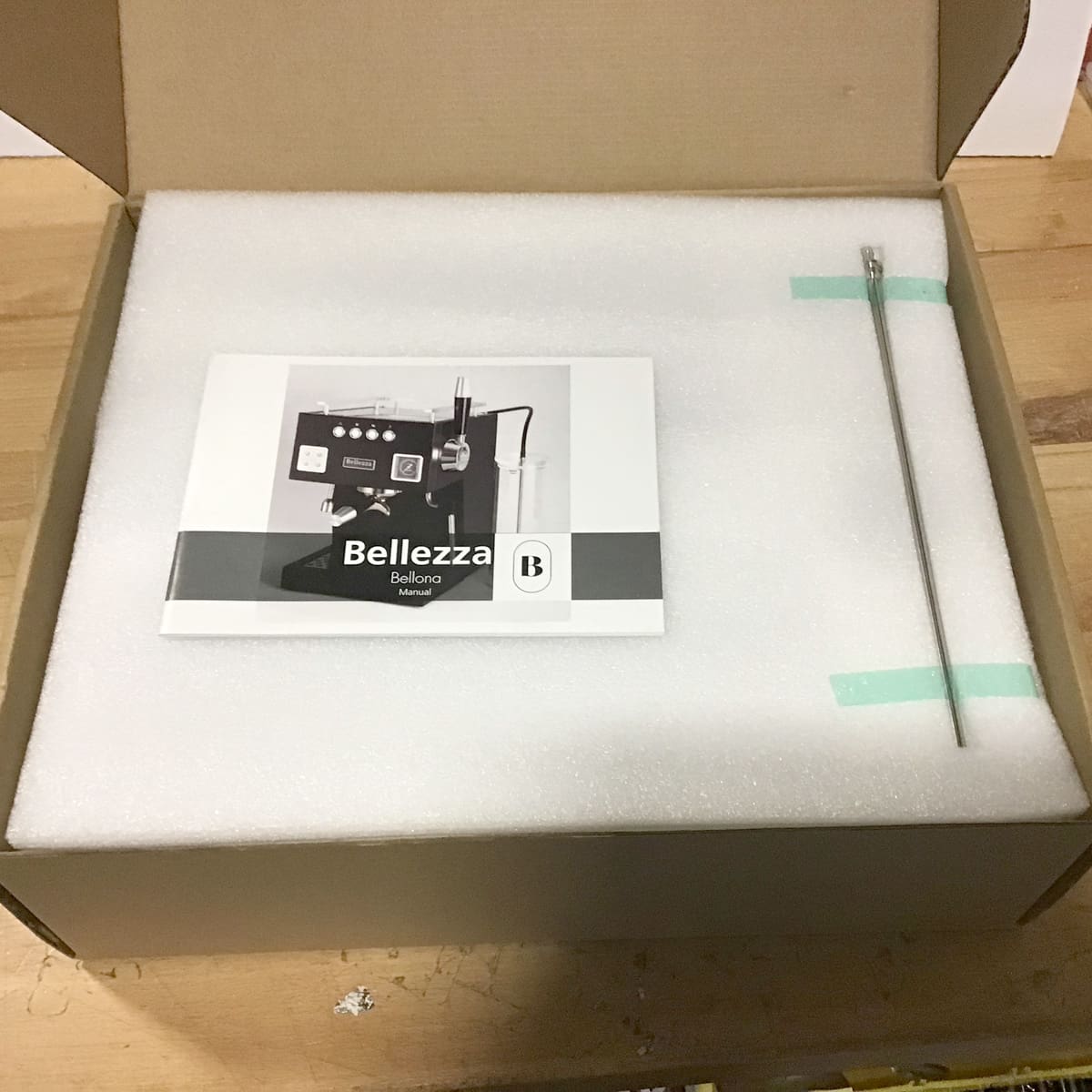 A Bellezza Bellona manual and metal rod rest on foam padding inside an open packing box, placed on a wooden surface. © 2025 1st-line.com