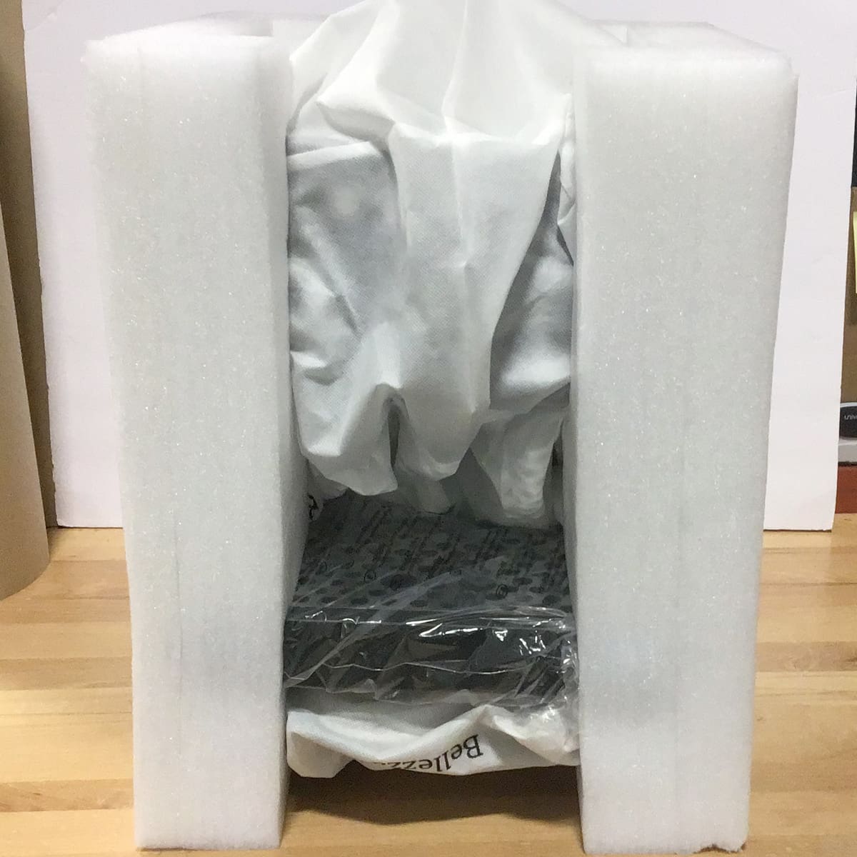 A Bellezza black electronic device wrapped in plastic and covered with white fabric is securely packed between two large white foam pieces on a wooden surface. © 2025 1st-line.com