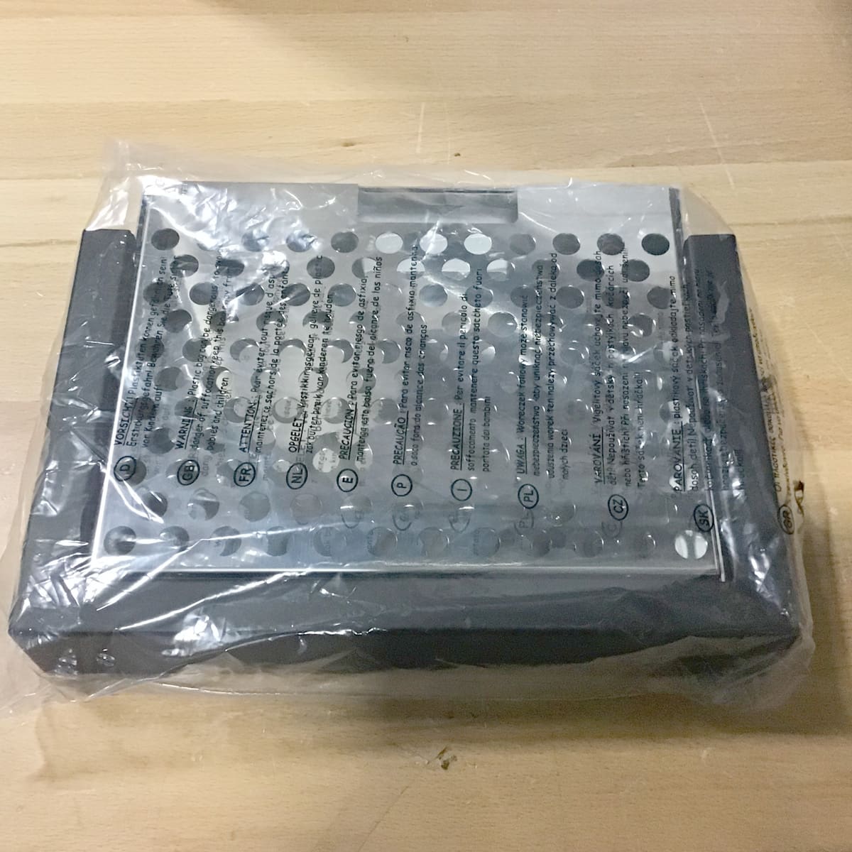 A Bellezza metal box grater with a black base is wrapped in clear packing and placed on a light wooden surface. © 2025 1st-line.com