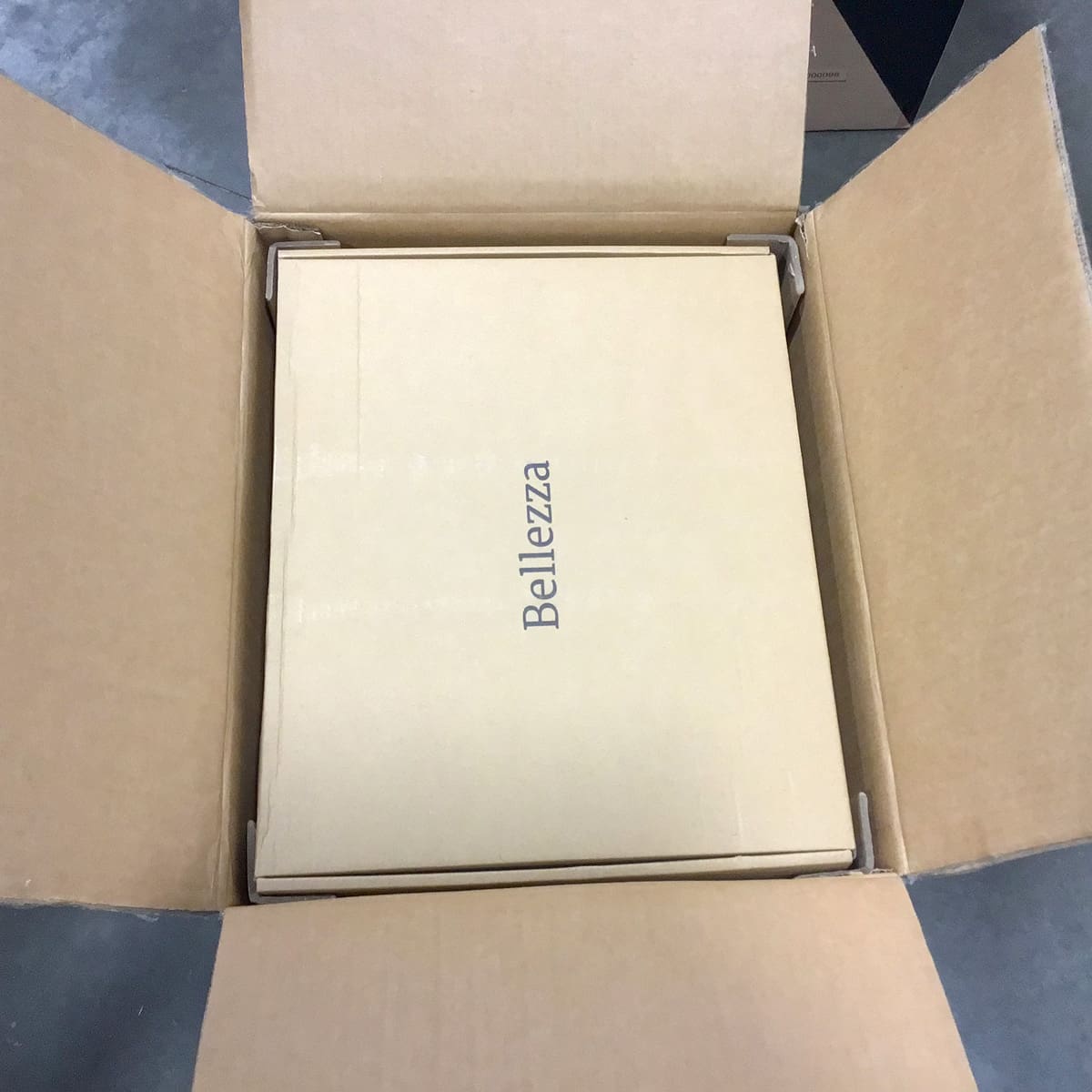 A cardboard packing box containing a smaller, flat box labeled Bellezza sits in the center on a concrete floor. © 2025 1st-line.com