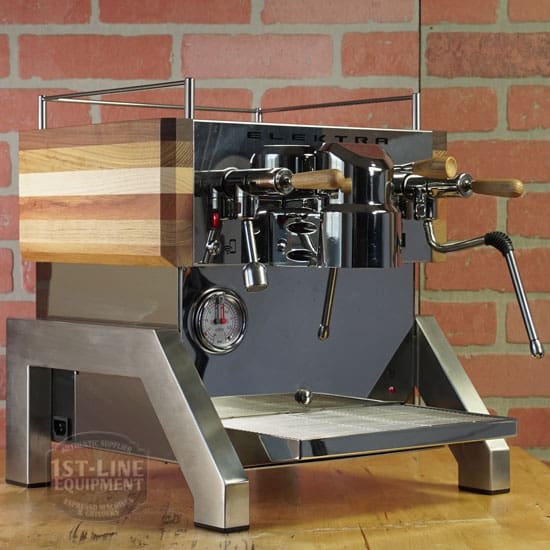 A LIKE NEW - Elektra Verve Dual Boiler Espresso Machine (C104) with polished steel and wood accents, dual levers, and a pressure gauge sits on a wooden countertop against a brick wall, highlighting its modern industrial design. © 2025 1st-line.com