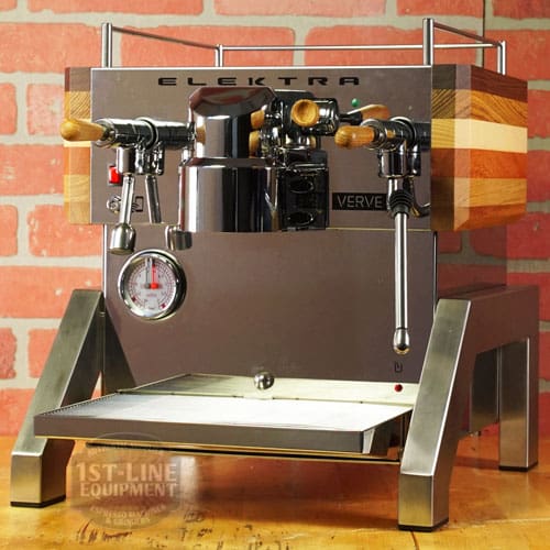 A LIKE NEW - Elektra Verve Dual Boiler Espresso Machine (C104) with stainless steel body, wooden handles and side panels, pressure gauge, steam wands, and drip tray sits against a brick wall background. © 2025 1st-line.com