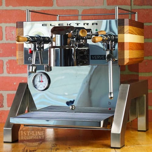A LIKE NEW - Elektra Verve Dual Boiler Espresso Machine (C104) with wooden handles and side panels sits on a counter against a brick wall, featuring a pressure gauge and steam wands on its stainless steel front. © 2025 1st-line.com