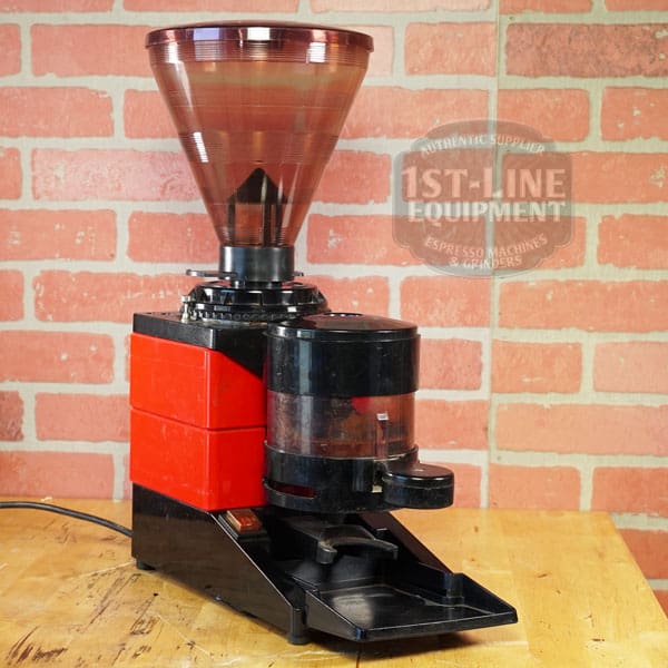 A red and black SIDEWALK SALE - Cunill Doser Grinder (E012) with a large bean hopper and grounds container sits on a wooden counter before a brick wall, featuring the 1st-Line Equipment logo—ideal for an E012 Sidewalk Sale display. © 2025 1st-line.com